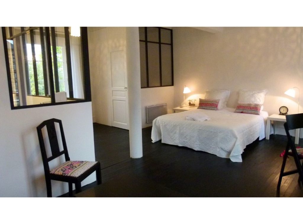 Suite nuptial dans un gîte.
Grande pièce blanche peinte en mat avec à gauche une salle de bains derrière une verrière. Cadre de la verrière et sol peints en noir satin. Joli mobilier simple et recyclé. Petite chaise noire année quarante avec une assise fleurie. Grand lit recouvert d'un joli bouti blanc. Petites lampe et plafonnier dentelle. Ambiance virginale, romantique, campagne nostalgique.