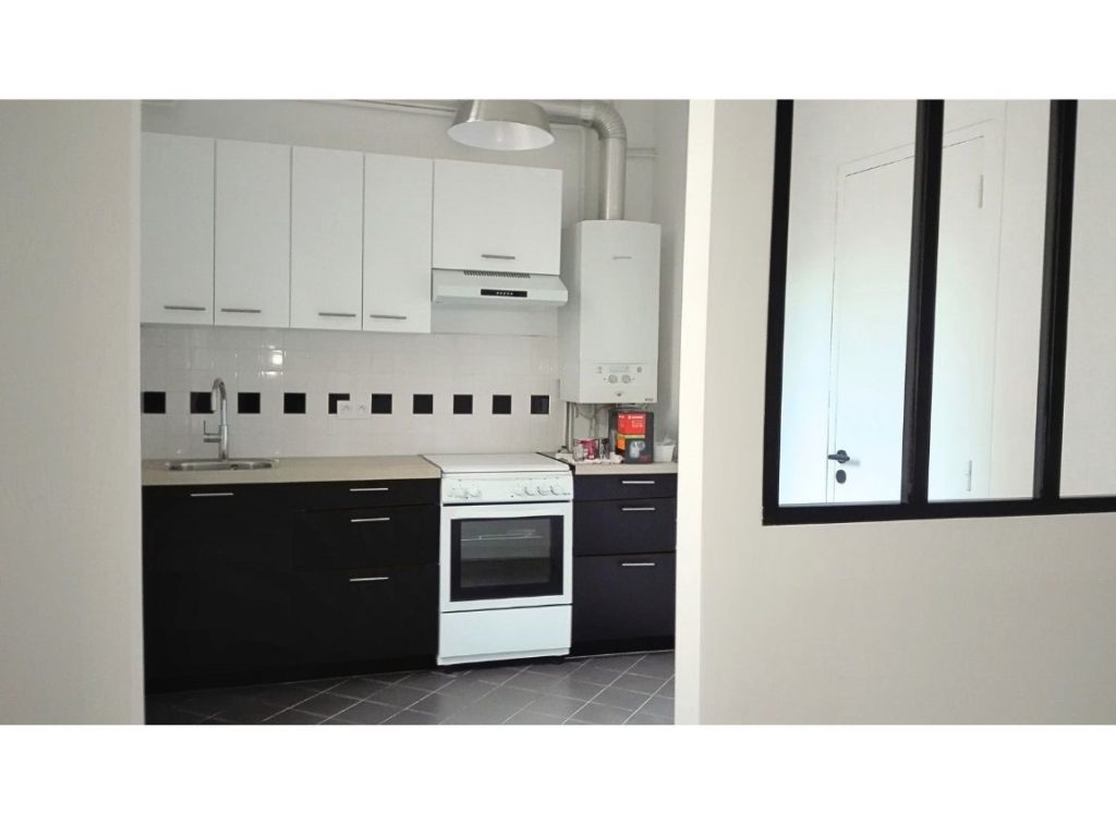 Cuisine en noire et blanc. Faîence de la crédence en petit carreaux blanc avec une frise de carreaux noirs. Les placard muraux sont blanc et ceux posés au sol sont noirs. Gazinière blanche. Murs blanc finition satin. On aperçoit la verrière entre la cuisine et le salon.