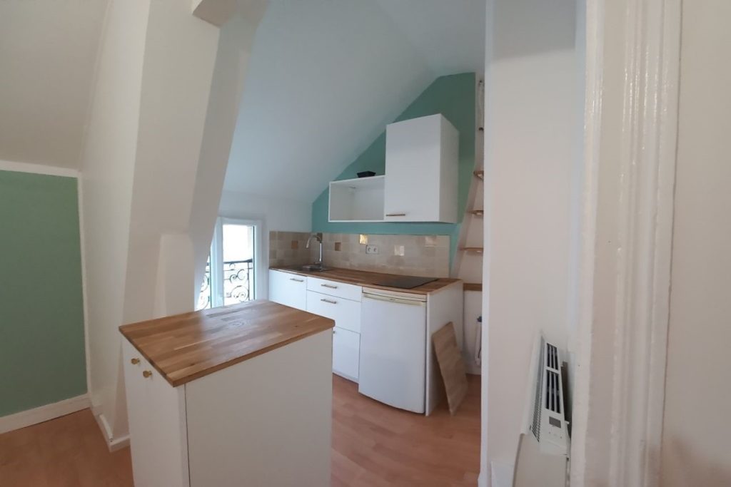 Cuisine lumineuse en sous pente. avec son petit ilôt centrale et un plan de travail en pin naturel traité. Frigo, évier, espace lave linge et des placards hauts dont un pour le micro-onde. Dominante couleur le blanc avec quelques murs en vert tilleul ou jade
