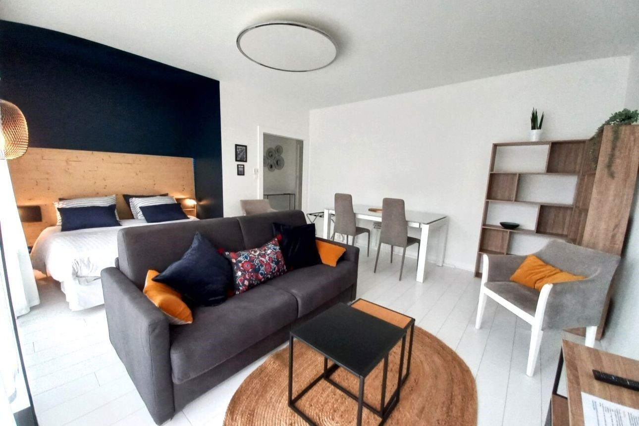 Rafraichissement peinture et réaménagement d'une grande pièce de vie dans un petit appartement de Paris. Salle de vie blanche avec un coin chambre délimité par une tête de lit en bois devant un mur bleu canard. En face du lit un espace salon TV avec un canapé gris, sur la droite le coin salle à manger avec son meuble bibliothèque.