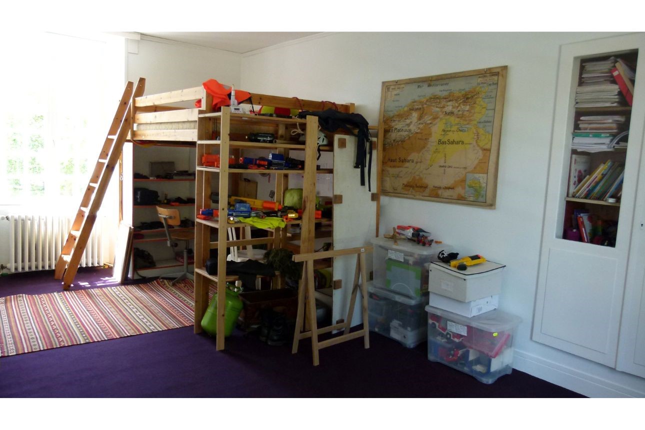 Chambre d'ado avant. Peinte en mat blanc avec sur un côté un lit en mezzanine avec un petit bureau dessous et plein de jouet entassés dans un coin, le tout sur une moquette violette.