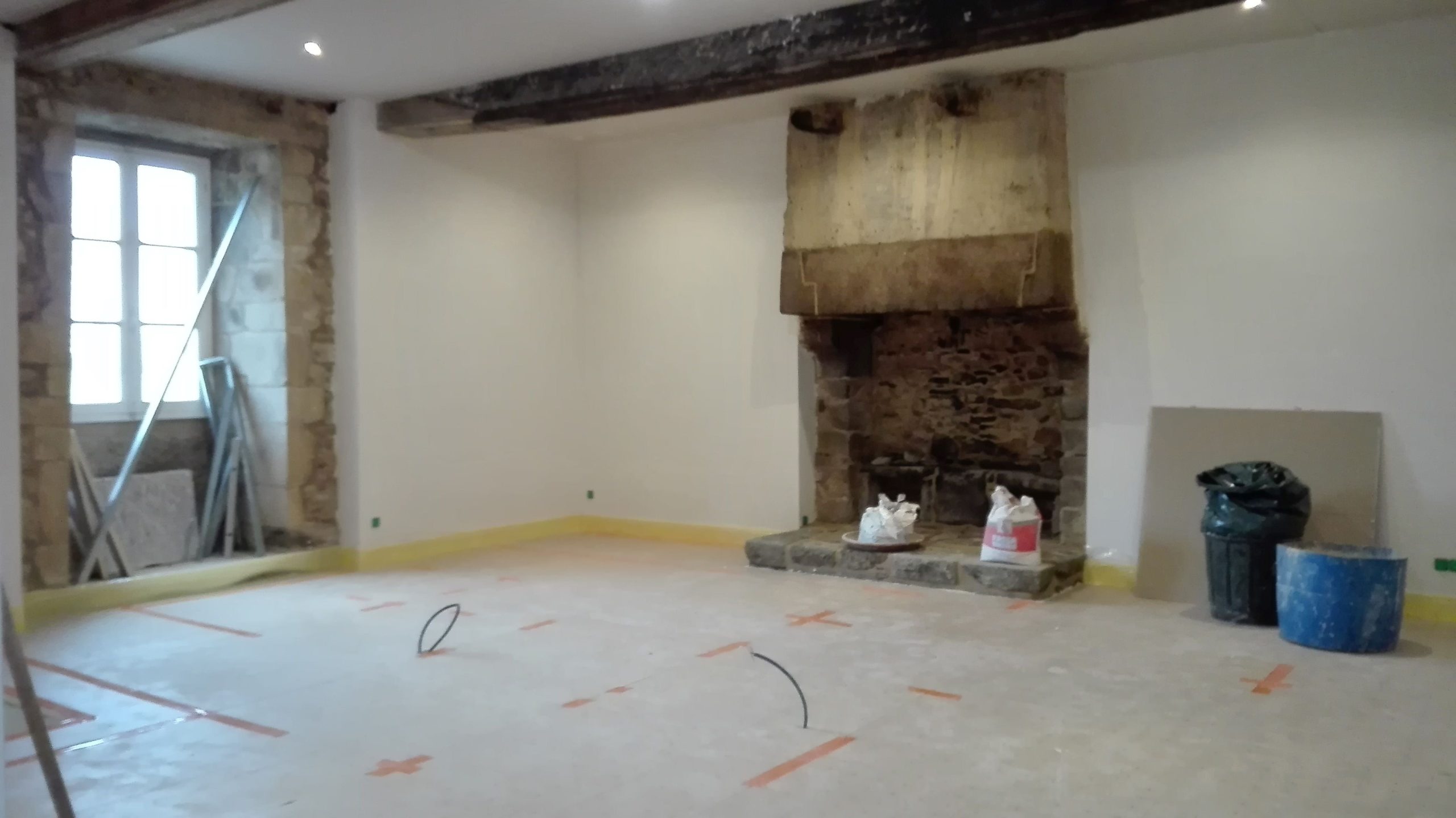 Mise en valeur d'une cheminée ancienne dans un manoir. Le manteau de celle-ci est en torchis de couleur ocre roux et le reste en granite beige. Les murs autour sont en plâtre blanc. Au-dessus sur le plafond blanc se détache de grosses poutres en chêne fumée marrons.